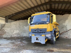 yellow gritting truck parked in a barn in front of a large pile of road salt,