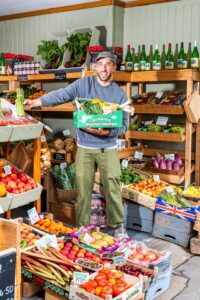 smiling man holding a box of veg in front of boxes of fruit and veg