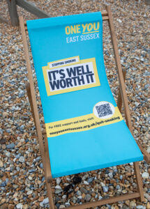 a turquoise deck chair on a shingle beach imprinted with the It's Well Worth It quit smoking message